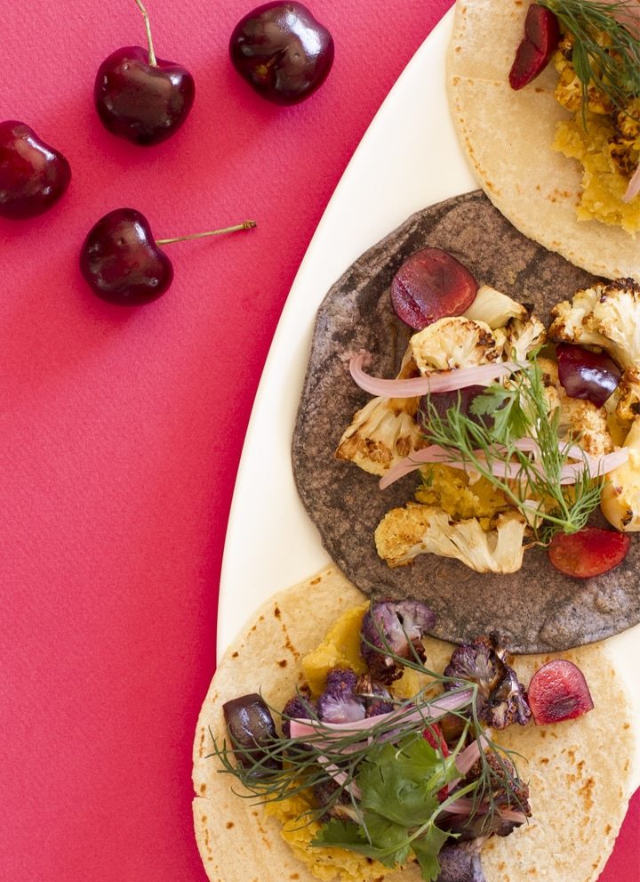 cauliflower, lentil, and cherry tacos featuring purple and yellow cauliflower over dal made from red lentils, topped with fresh cut cherries, pickled red onions, fresh dill and cilantro over white and purple corn tortillas on a white plate with a magenta background.