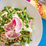 Asparagus ribbon salad with sliced sugar snap peas, sliced pink watermelon radish, cooked white beans, and grated white cheddar cheese on a large, round, white plate over a blue surface. Tie-dye yellow, pink, and white dotted napkin with forks shown in the upper right.