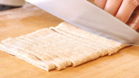 A hand holding a chefs knife and slicing rolled and folded whole wheat undo noodle dough on a wood cutting board.