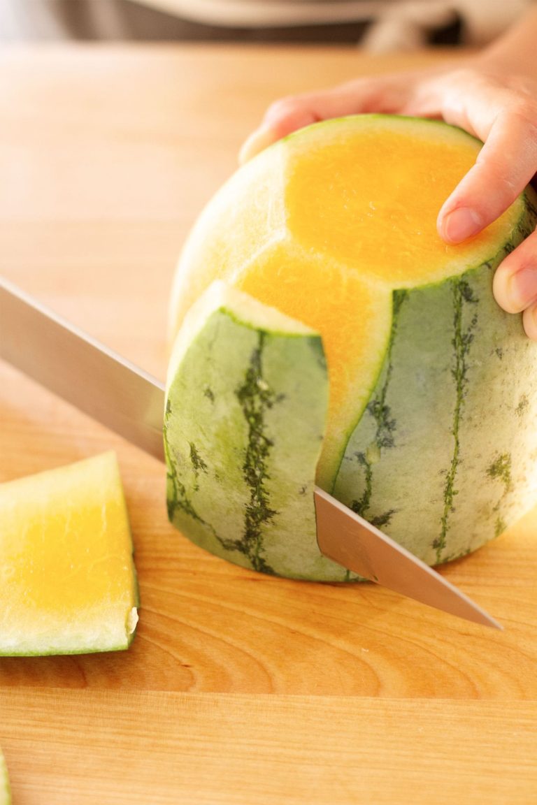close up of knife shaving the skin off of a halved orange colored watermelon