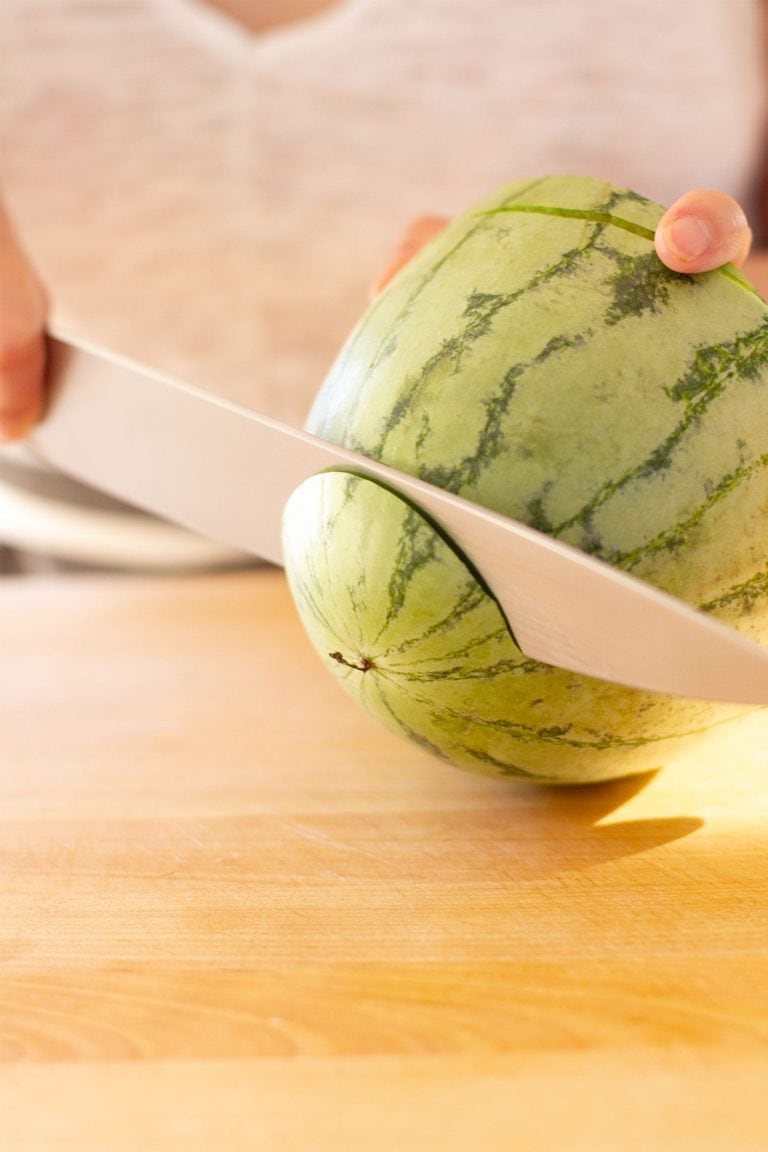woman in white cutting the tops off a halved watermelon on cutting board.