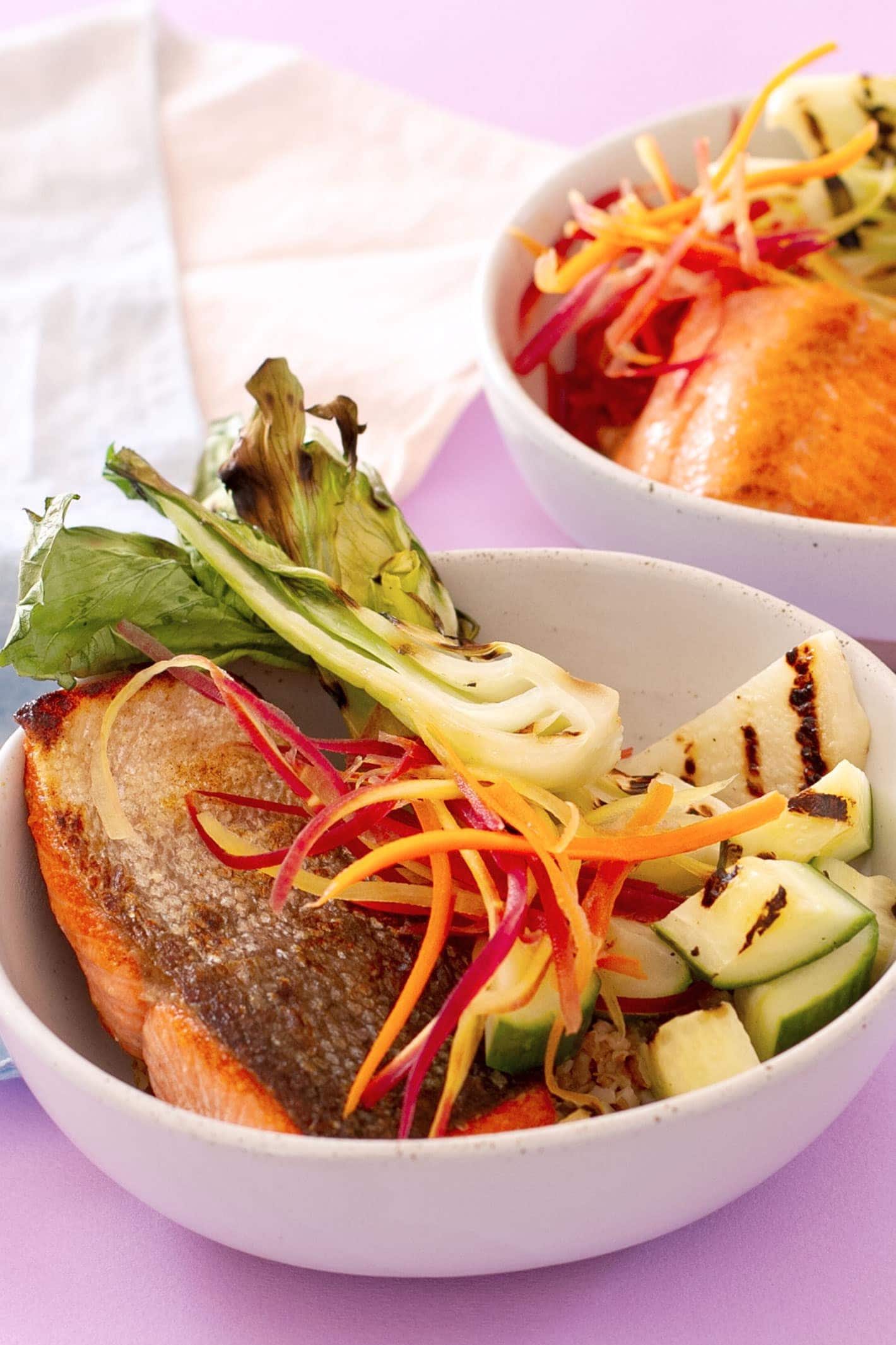 Close-up view of a grain bowl with pan-seared salmon, carrot strips, grilled baby bok choy, kohlrabi, and cucumber over a bed of bulgur in a white bowl.