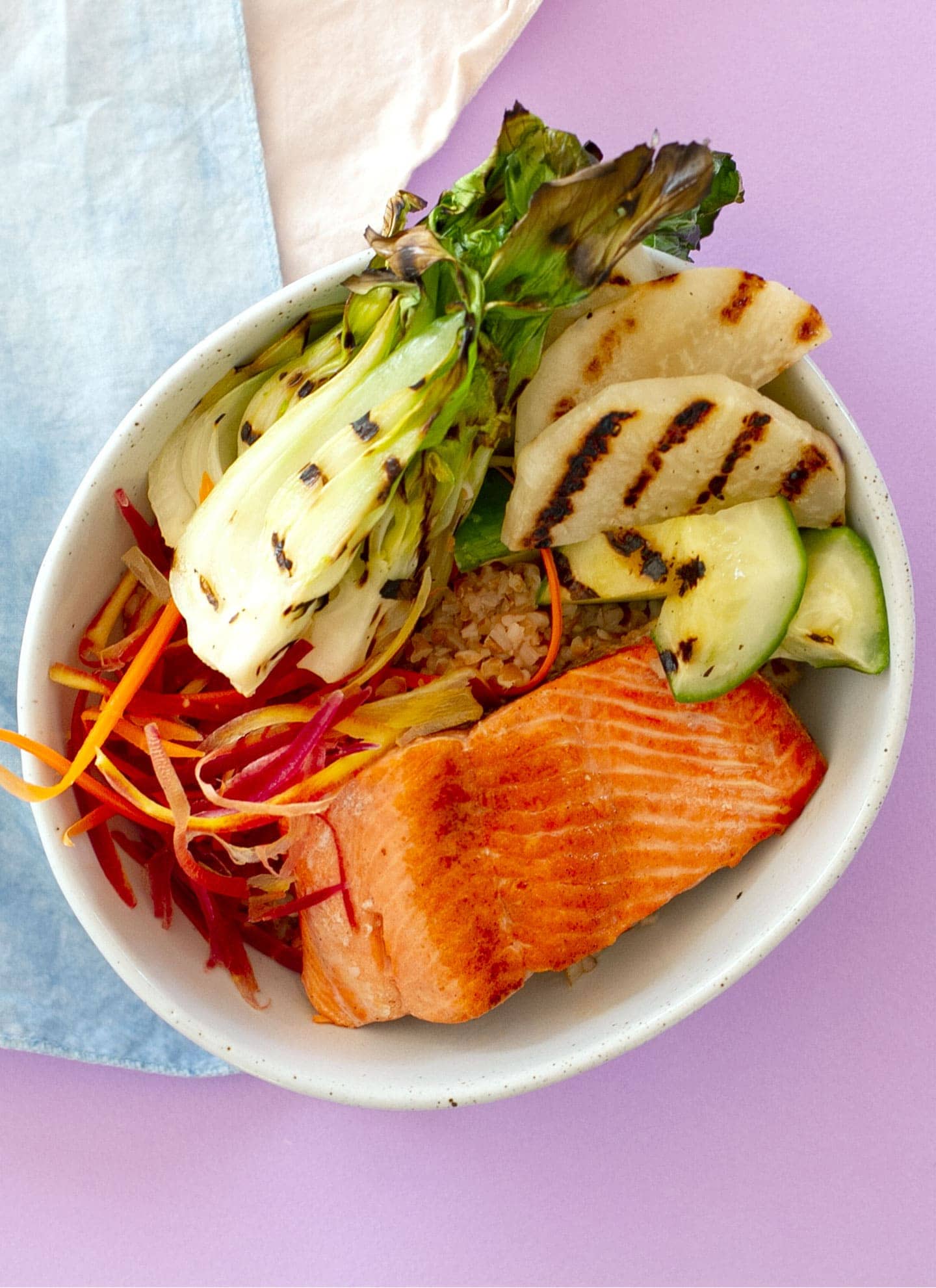 Top-down view of a grain bowl with pan-seared salmon, carrot strips, grilled baby bok choy, kohlrabi, and cucumber over a bed of bulgur in a white bowl sitting on a pastel purple surface.