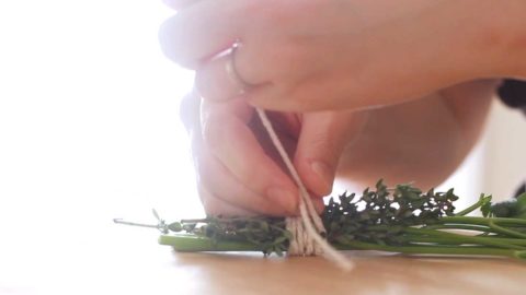 Two hands tying kitchen string around a bouquet of fresh thyme and parsley stems.