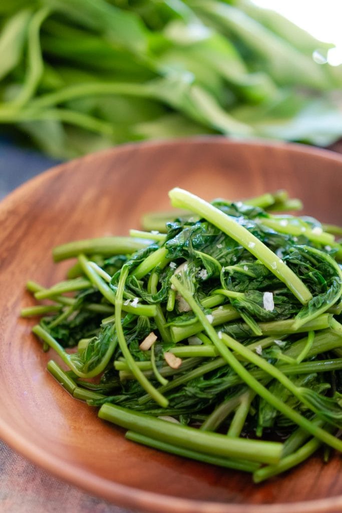 A close up view of stir-fried water spinach seasoned with flaky salt on a wood plate.