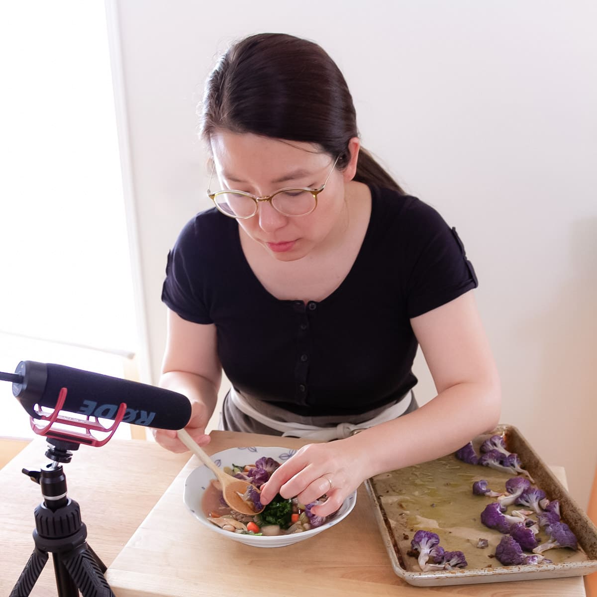 Cindy food styling a bowl of colorful chicken and vegetable soup with a tray of purple cauliflower on the side and a Rode microphone in the front.