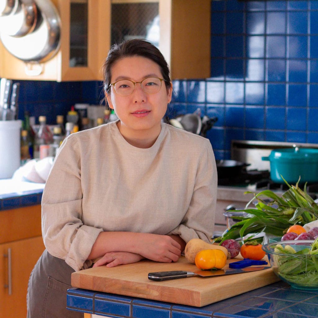 Cindy smiling while wearing a beige shirt and gray apron, leaning on a cutting board surrounded by fall produce.