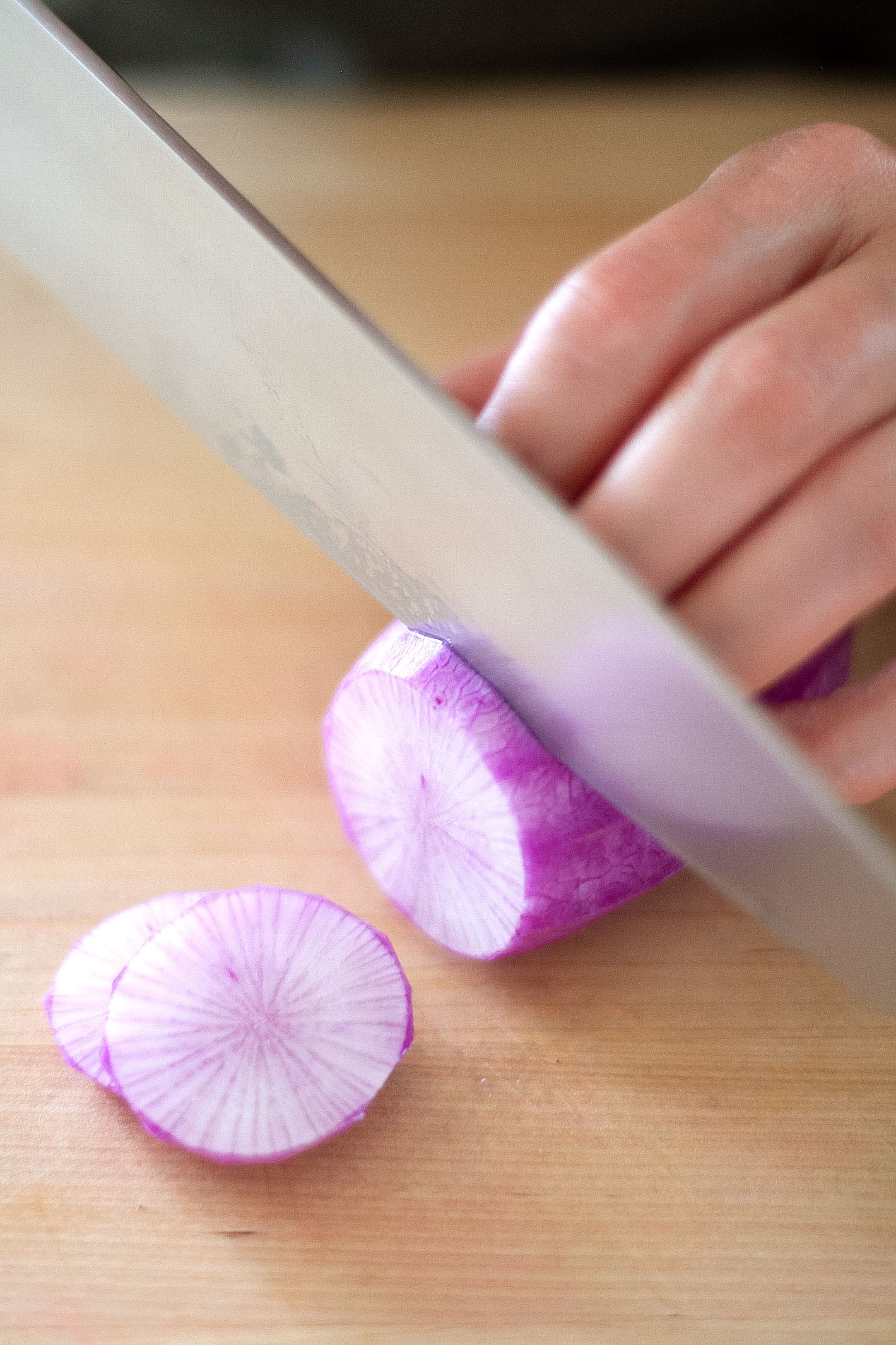 A hand hold peeled purple daikon radish on a cutting board with a chefs knife cutting slices.